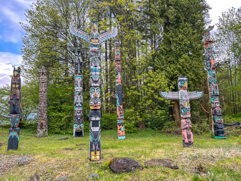 many totem poles standing on grassy hill with trees behind and sky above.
