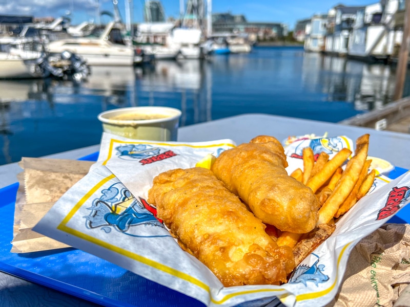 two pieces of crispy fish and chips in basket on table with boats and water behind.