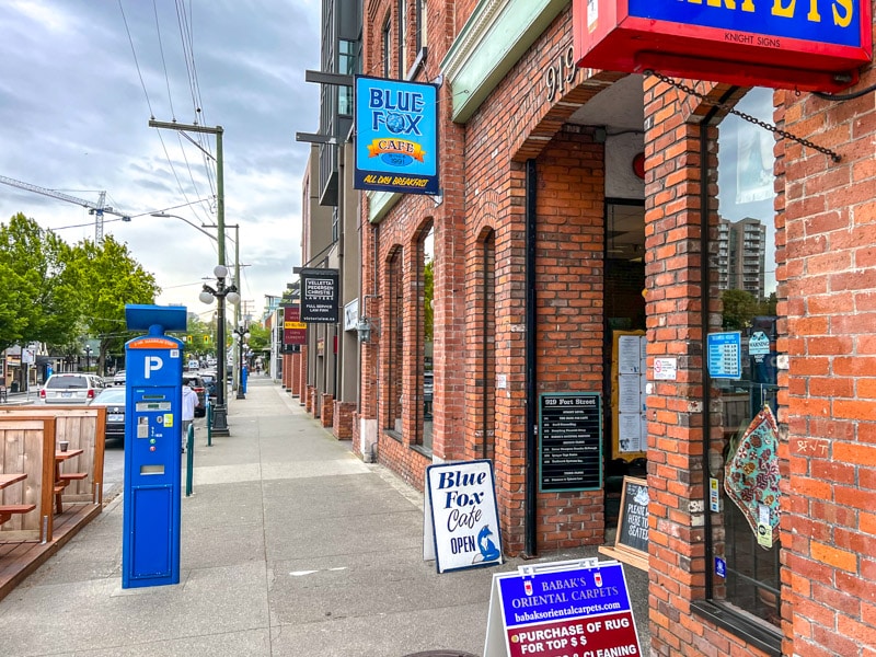 Blaues Schild für ein Café, das an einer roten Backsteinmauer über dem Bürgersteig in Victoria B.C. hängt.