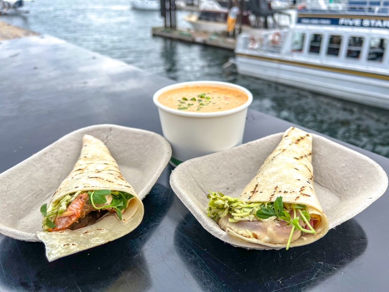 two hand rolled fish tacos and a bowl of chowder on table with harbour water and boats behind.