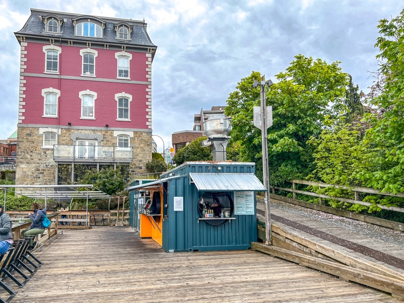 small shipping container on wooden boardwalk with red building behind on hillside.