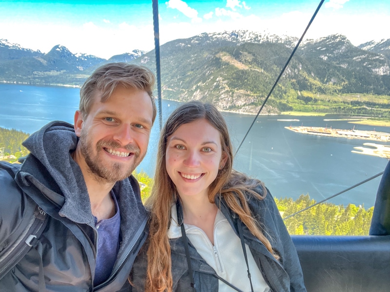 man and woman taking selfie in green gondola with water and mountains behind.