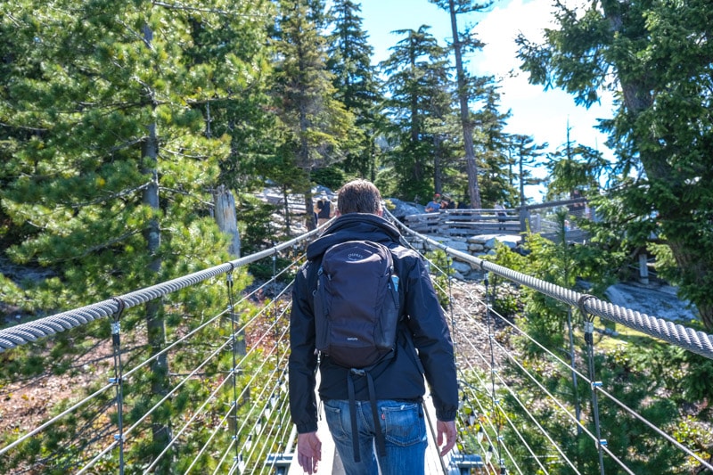 back of man wearing jacket and backpack crossing suspension bridge.