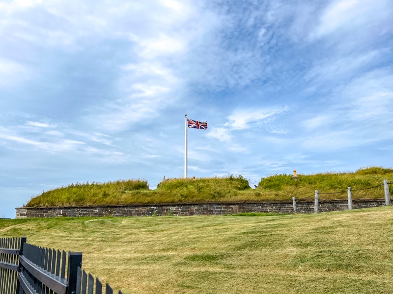 union jack flag on white pole flying above green grassy walls of fort with sky above.