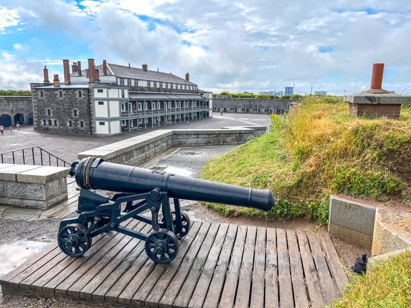 large black cannon on top of citadel wall with large building and open square in background.