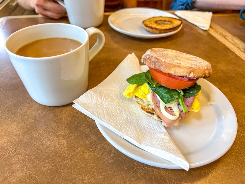 small breakfast sandwich on white plate with large coffee in mug beside on brown cafe table.