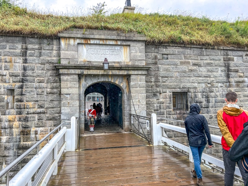 stone entrance to citadel with wooden bridge in front and red coat guard standing still.