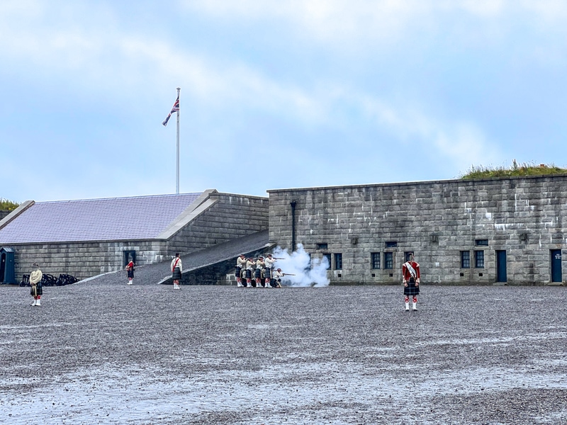 small troop shooting rifles at stone wall inside historic fort with sky and flag pole above.