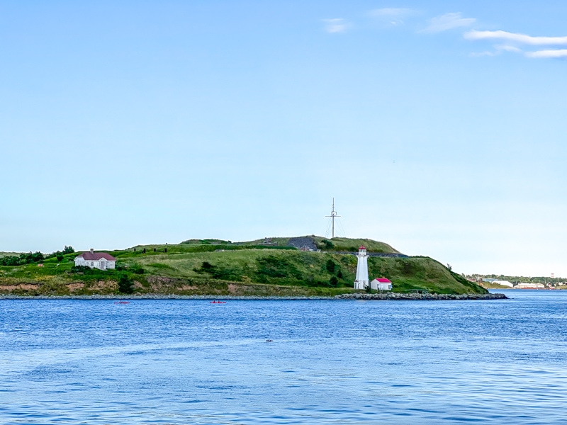 green island with small white lighthouse surrounded by harbour water and blue sky above.