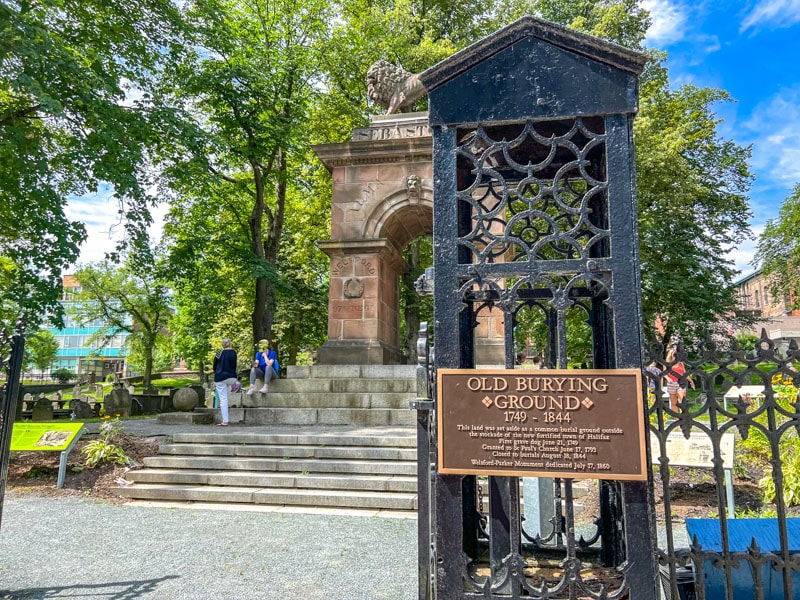 Metallschild auf schwarzem Torpfosten mit Friedhof und Denkmal im Hintergrund.