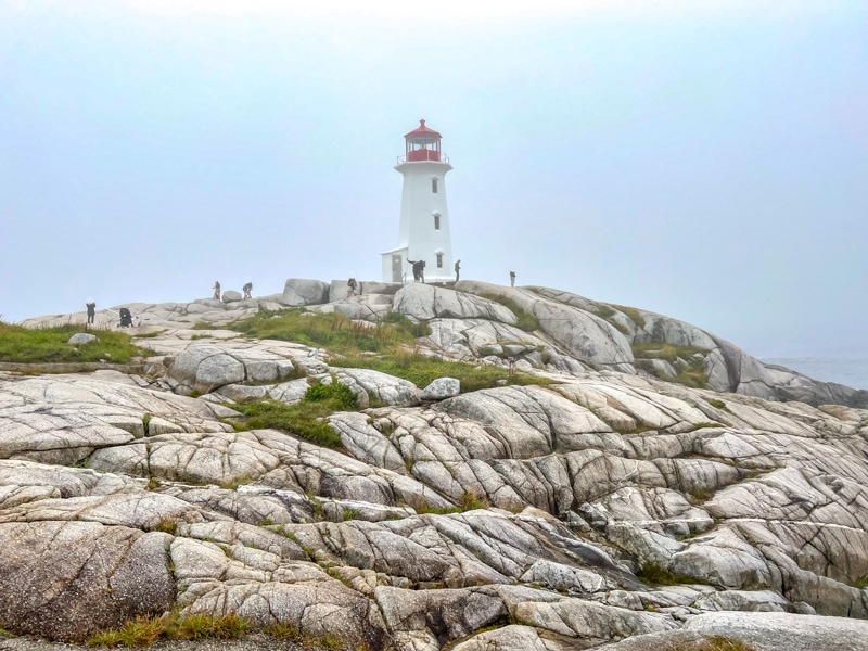 foggy shot of white and red lighthouse at peggys cove with rocky landscape around and people walking on them.
