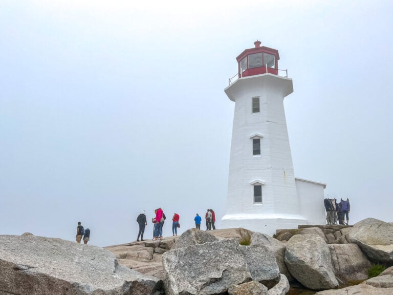 white lighthouse with red top sitting on rocky shoreline with people walking around under gloomy skies.