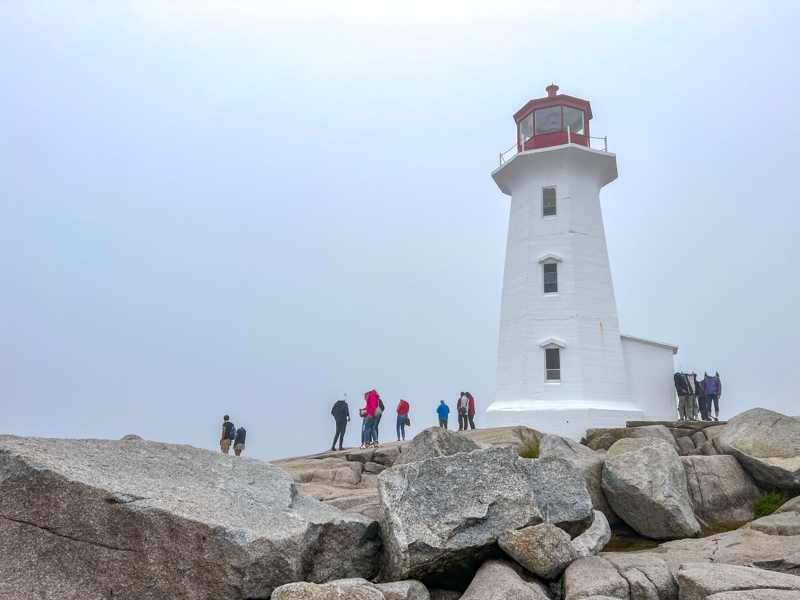 white lighthouse with people standing at the base with large rocks around and grey sky above.