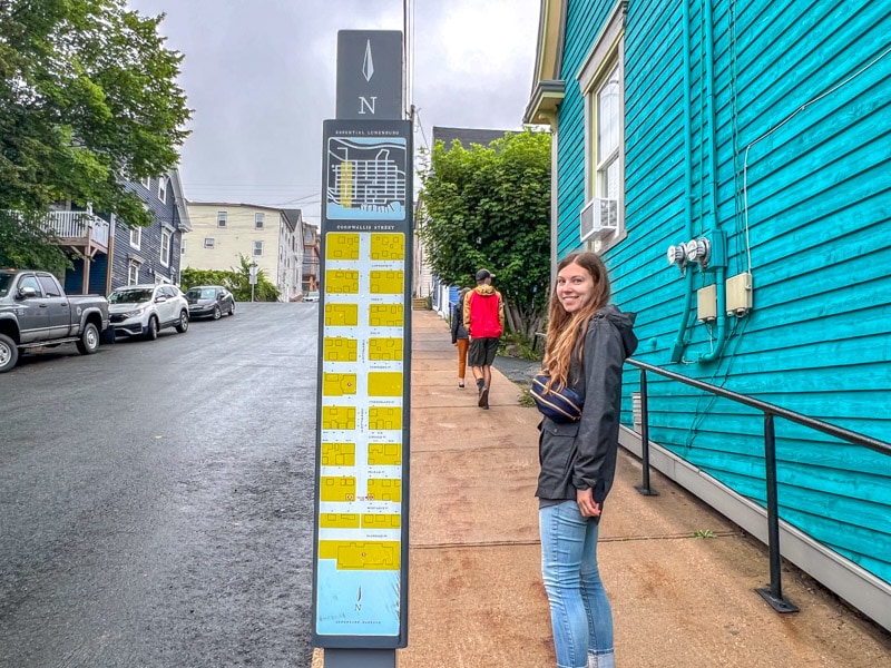 Frau steht neben Straßenschild auf dem Hügel mit blauem Haus daneben.