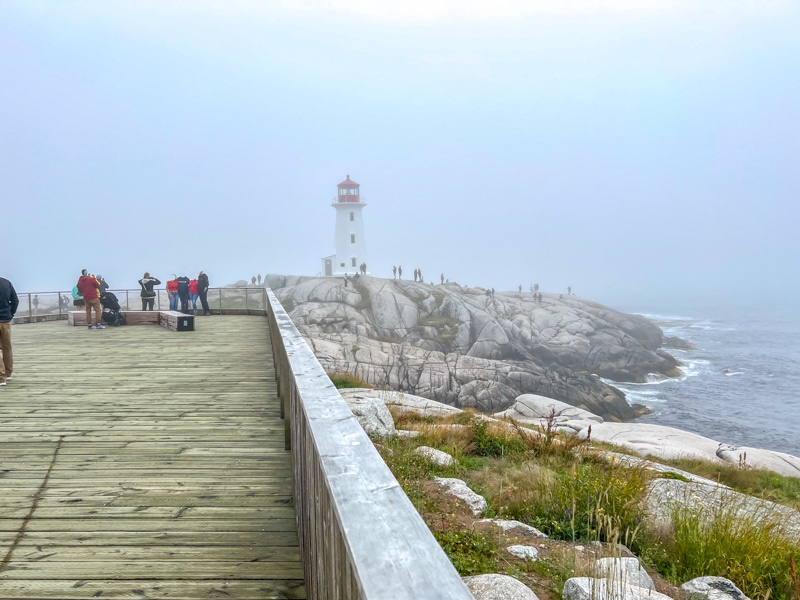 Holzpromenade mit Leuchtturm und felsiger Küste im Hintergrund.