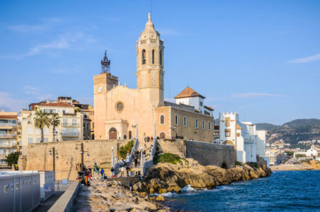 church with tower sitting atop rocky shoreline in sitges spain with water below.