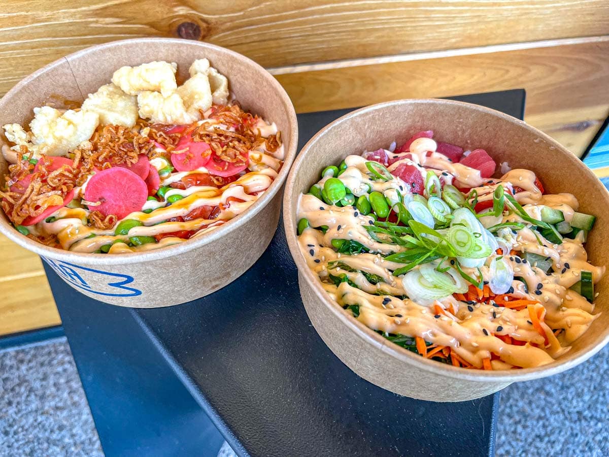 two poke bowls with los of ingredients sitting on small table with wooden all behind.