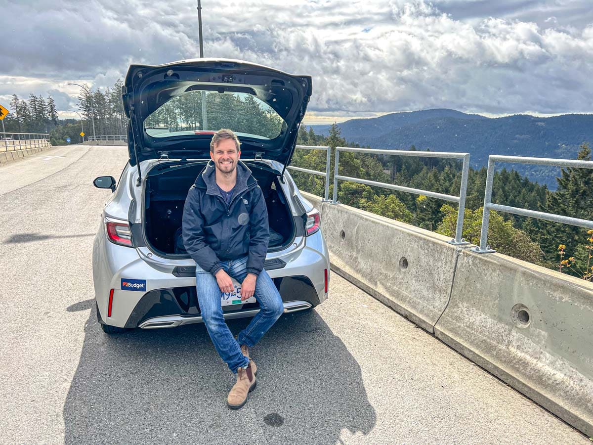 man sitting in trunk of silver car at rest stop with mountains in distance on vancouver island.