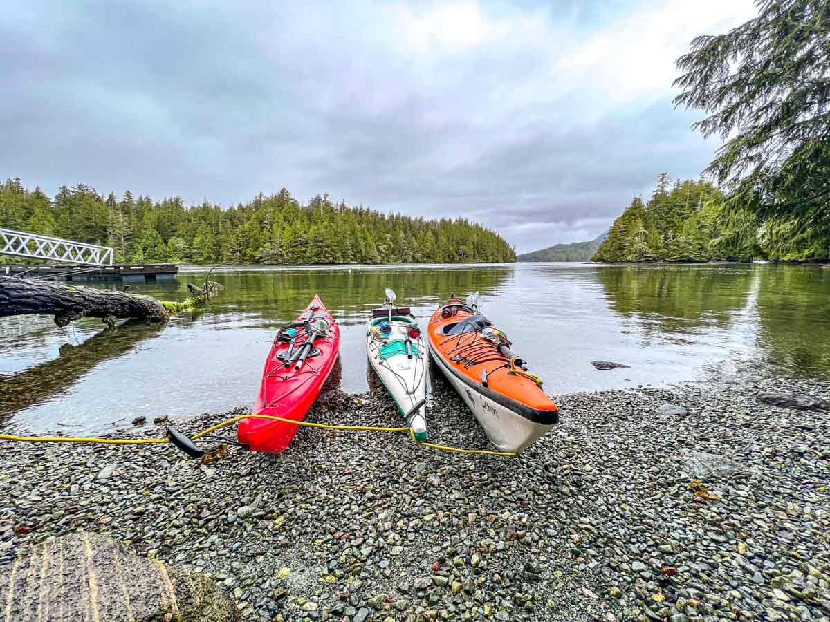 three colourful sea kayaks tied together on beach with water and trees behind.