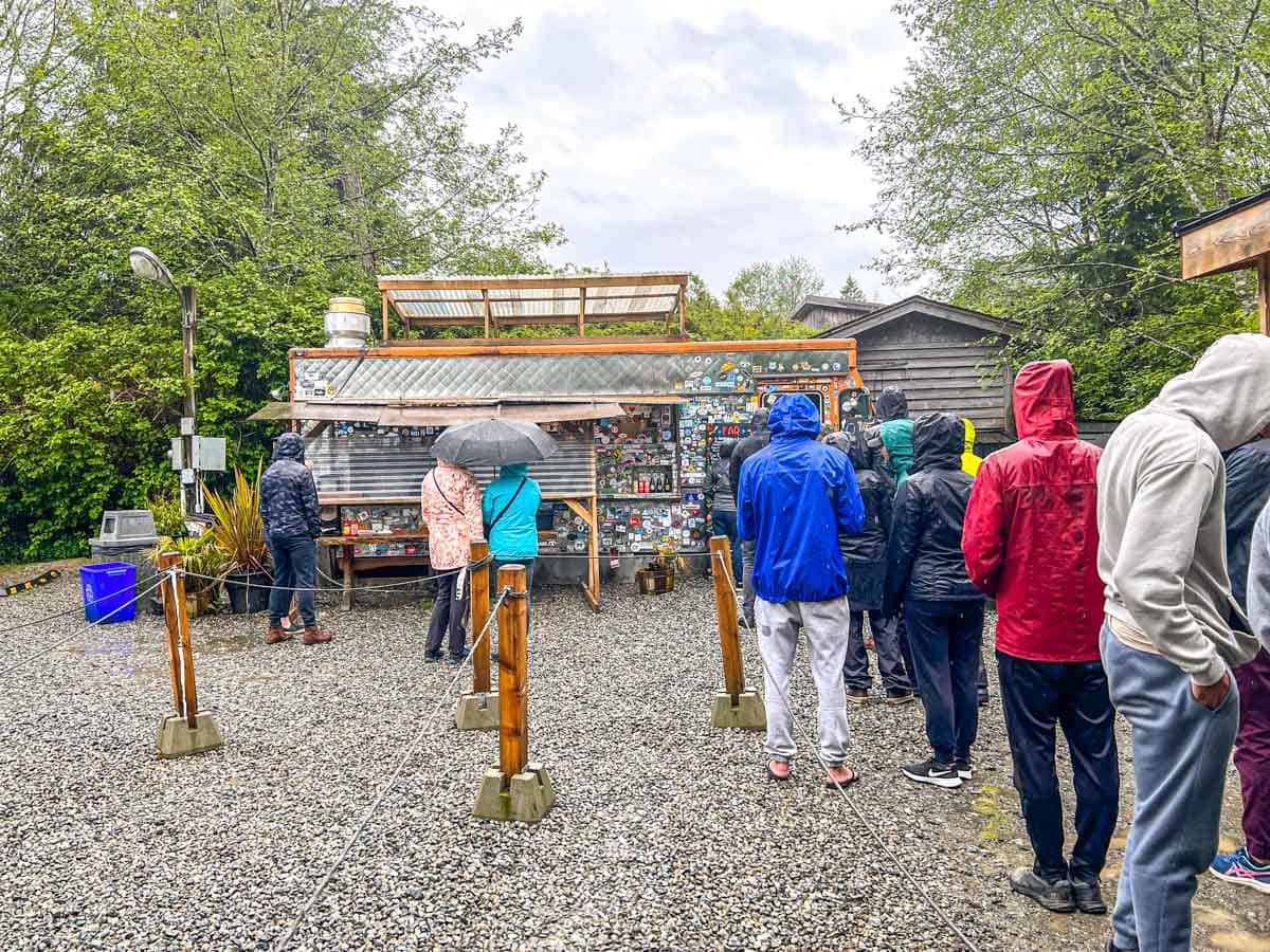 people lined up in the rain for orange food truck with sky and trees behind.