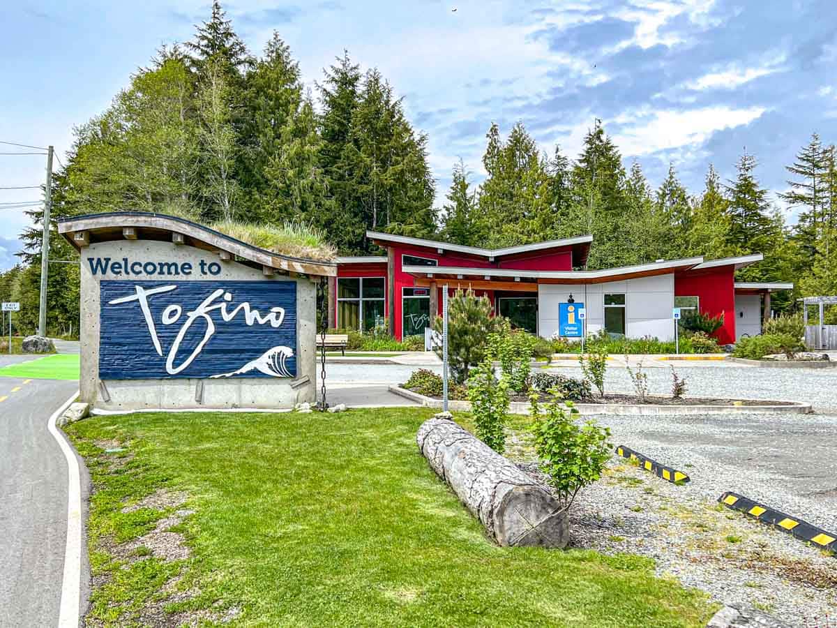 red building behind large blue welcome sign for tofino bc with trees behind.