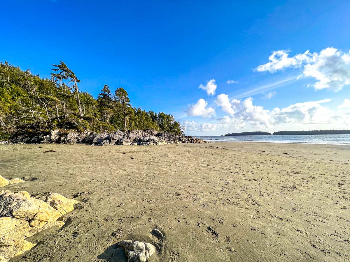sandy beach with blue sky above green trees and rocky formations.