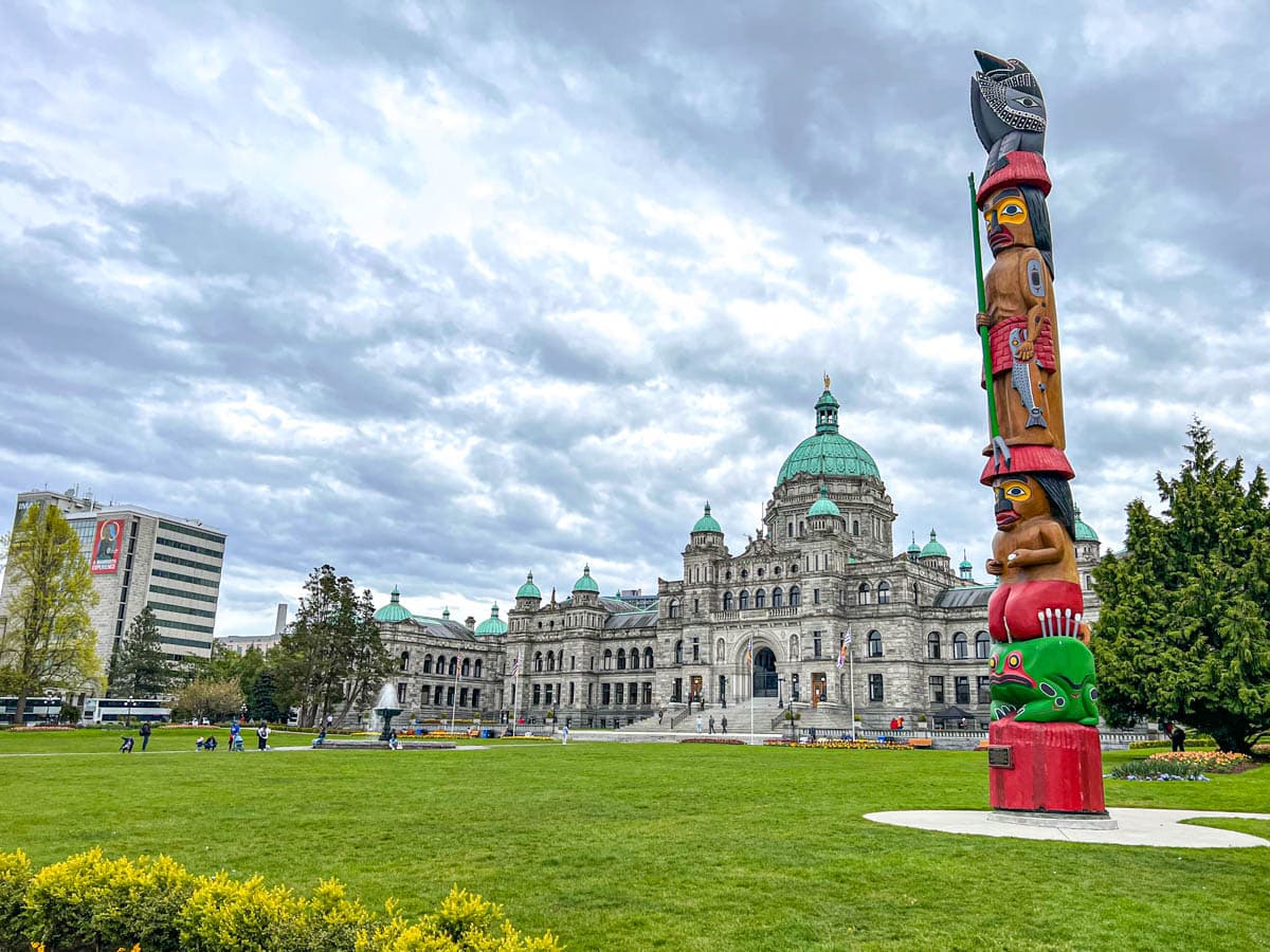 large building with green dome and green lawn behind colourful totem pole in foreground in victoria bc.