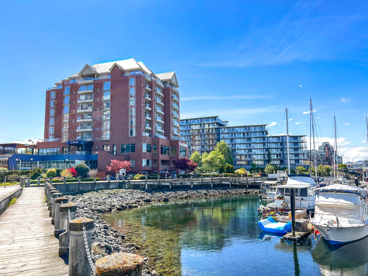 Ein großes Hotel aus rotem Backstein steht neben einer hölzernen Promenade mit Segelbooten im Hafen von Victoria.