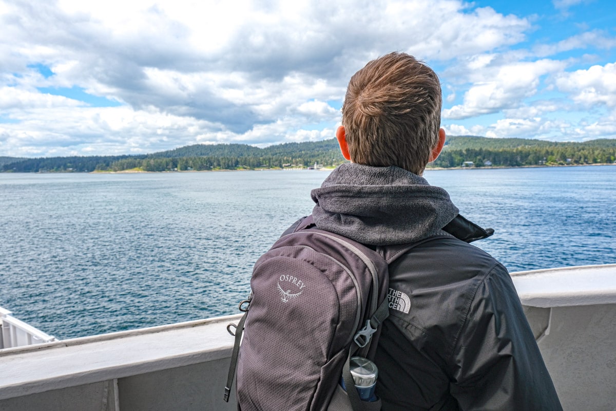 man wearing jacket and backpack staring out over sea from ferry.