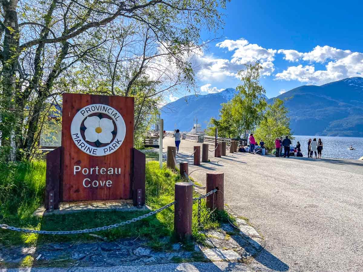 wooden sign for porteau cove with coastline and mountains in distance behind.