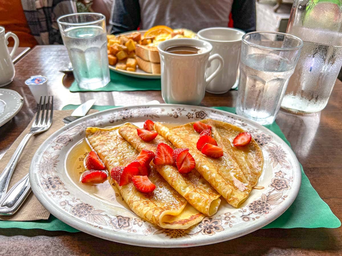 plate of crepes with syrup and strawberries on table with coffee mugs.