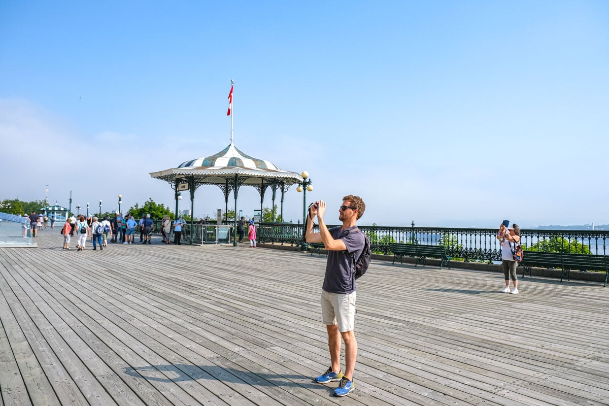 man taking photo of something standing on wooden terrace with blue sky behind.