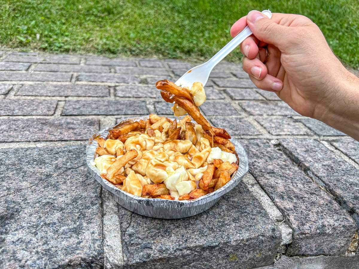 hand holding fork pulling fries and cheese out of a plate of poutine on rocky ledge.