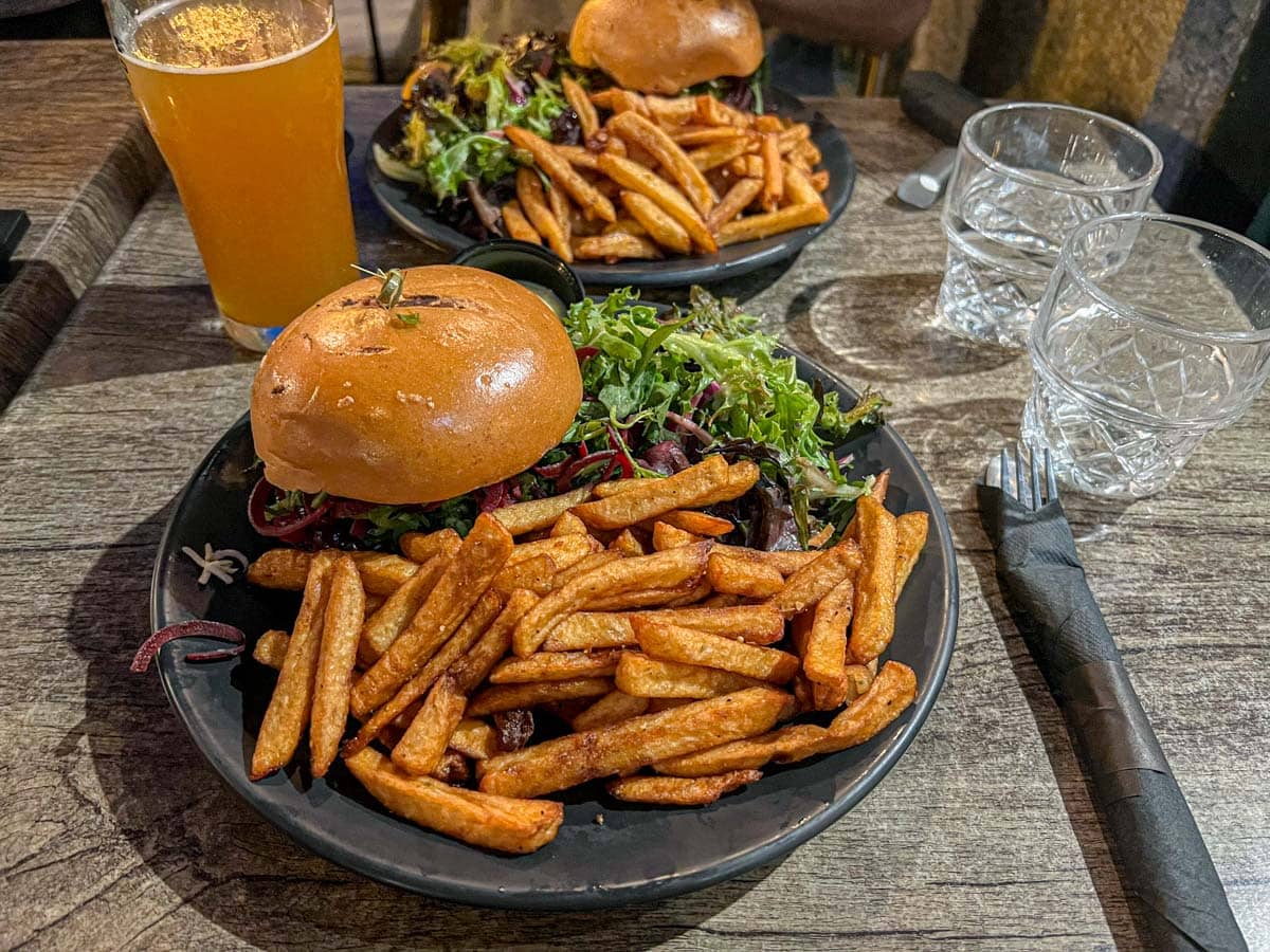 two burgers on plates with fries and a pint of beer on the table beside.