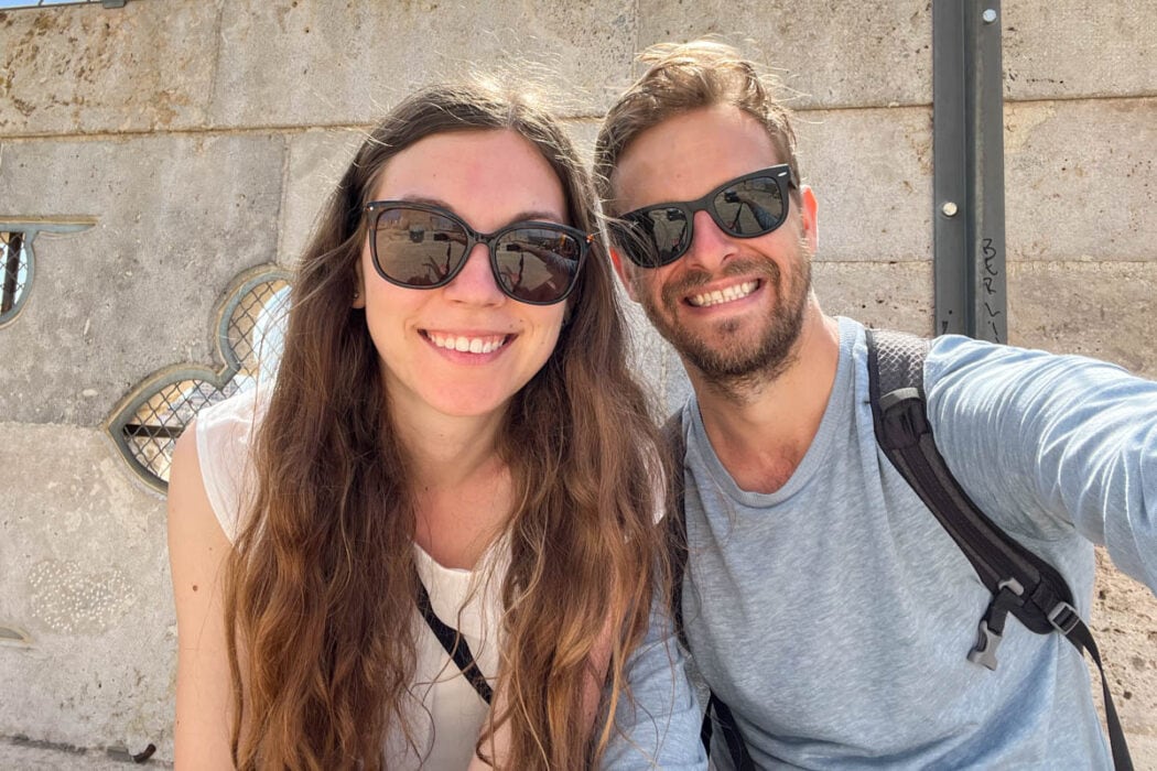man and woman both wearing dark sunglasses smiling for  camera with stone wall behind.